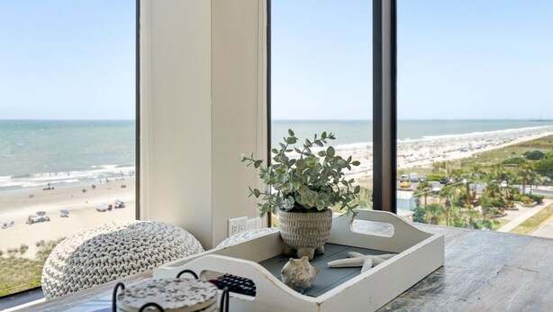 Habitació amb una finestra gran que dona a una platja, una planta en test, un pouf blanc i un comandament a un safata.