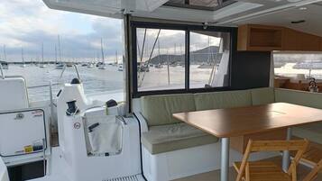 Interior de un barco con comedor, una pequeña mesa y vista al puerto deportivo.