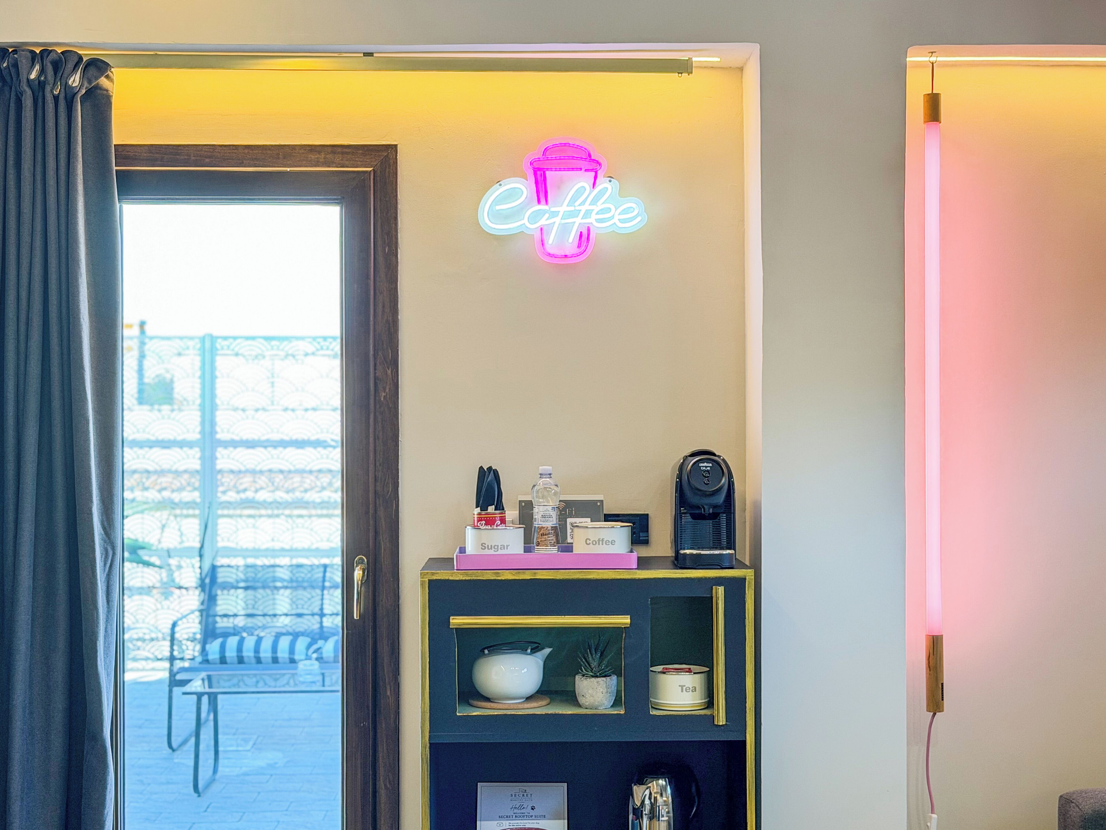 A room with a neon sign reading 'Coffee' above a shelf with a coffee machine, sugar, and a tea container.