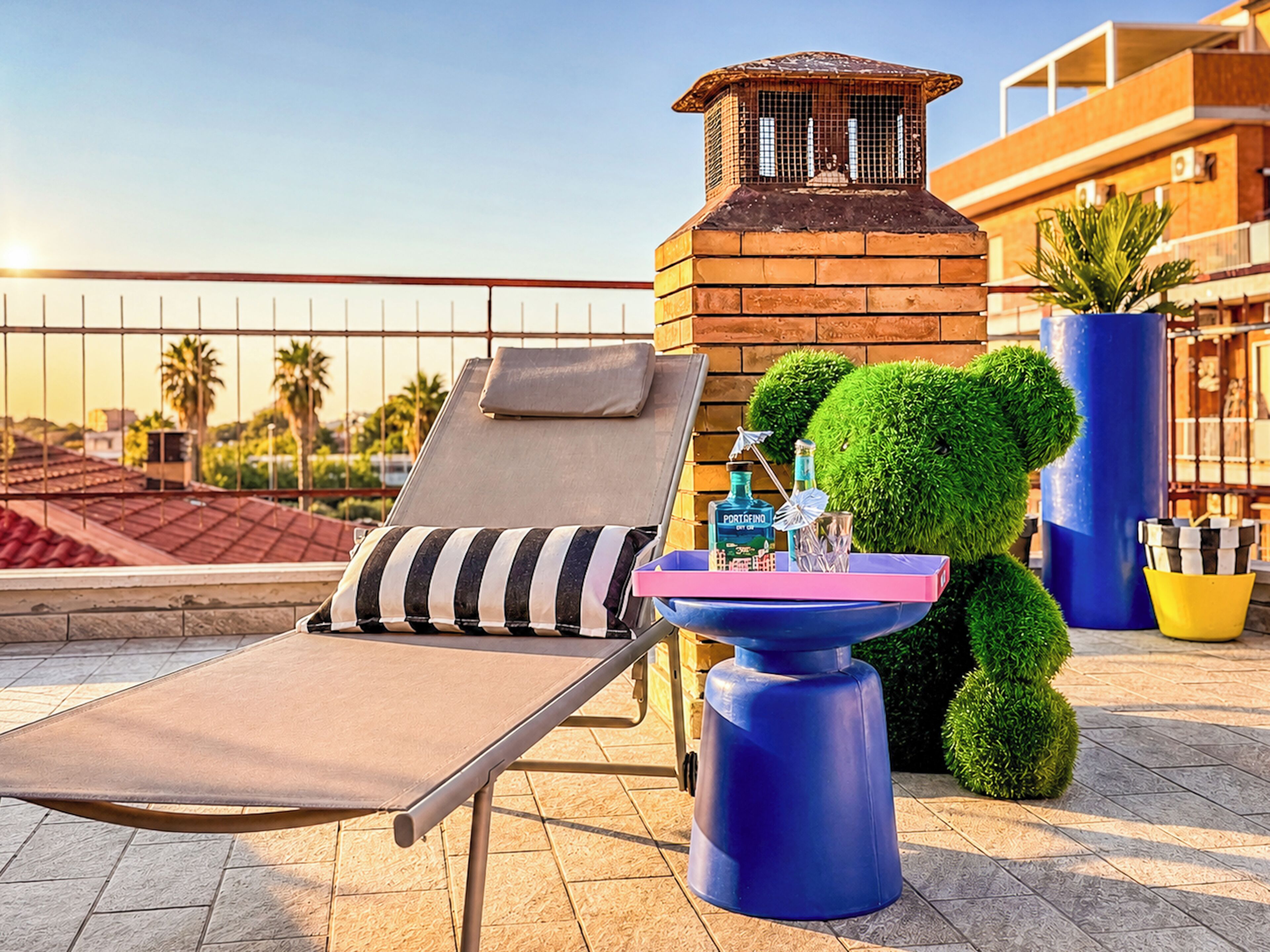 Un salón en la azotea con un sillón de sol, una pequeña mesa con una botella y un paraguas, y un árbol decorativo.
