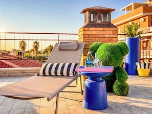 Un salón en la azotea con un sillón de sol, una pequeña mesa con una botella y un paraguas, y un árbol decorativo.