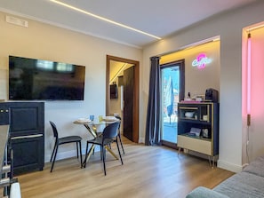 Una habitación moderna con televisión de pantalla plana, una zona de comedor con mesa y sillas de vidrio, una estación de café y vistas a través de una puerta.