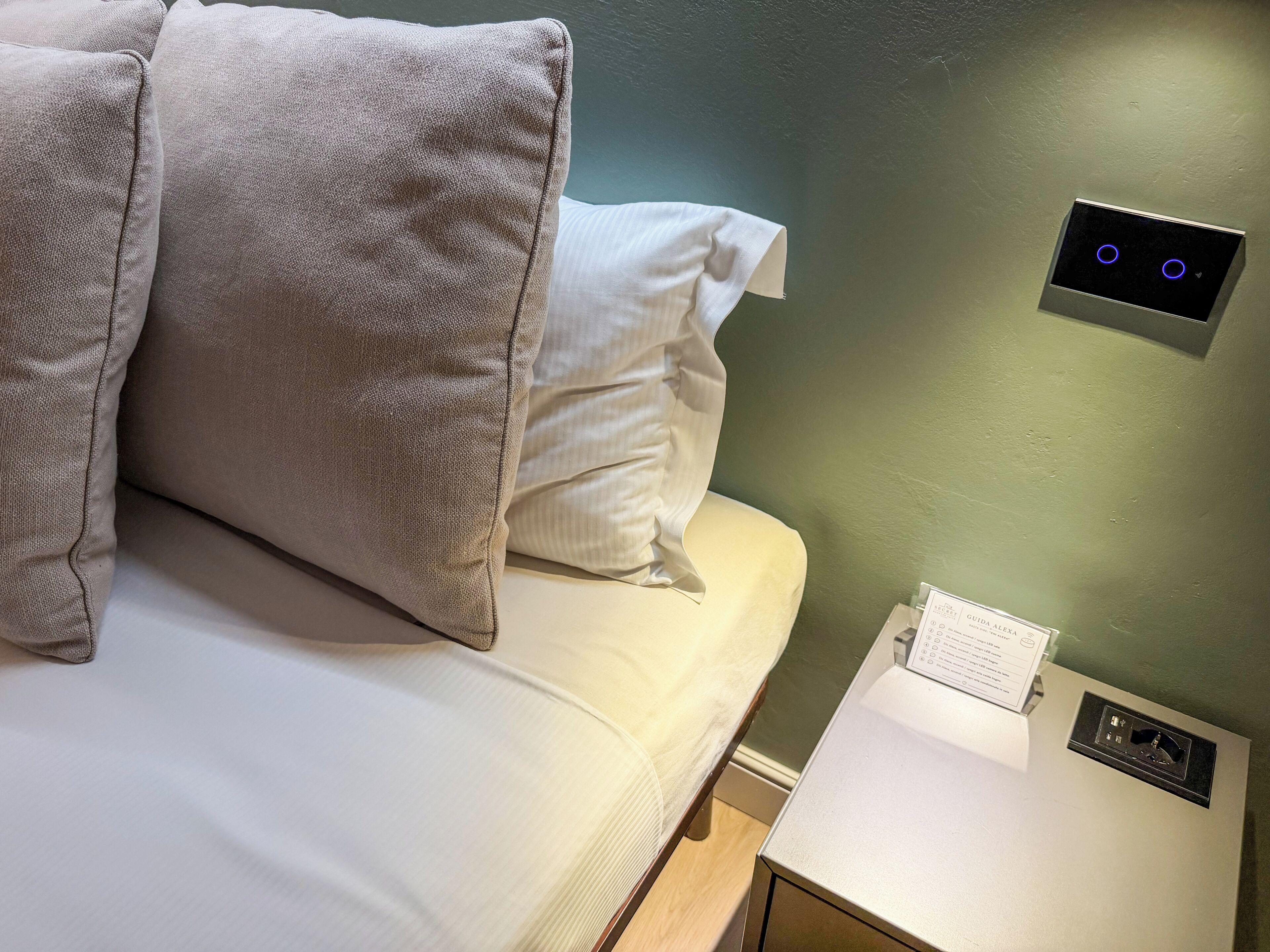 A hotel room with a bed, pillows, a nightstand, and a wall control panel.