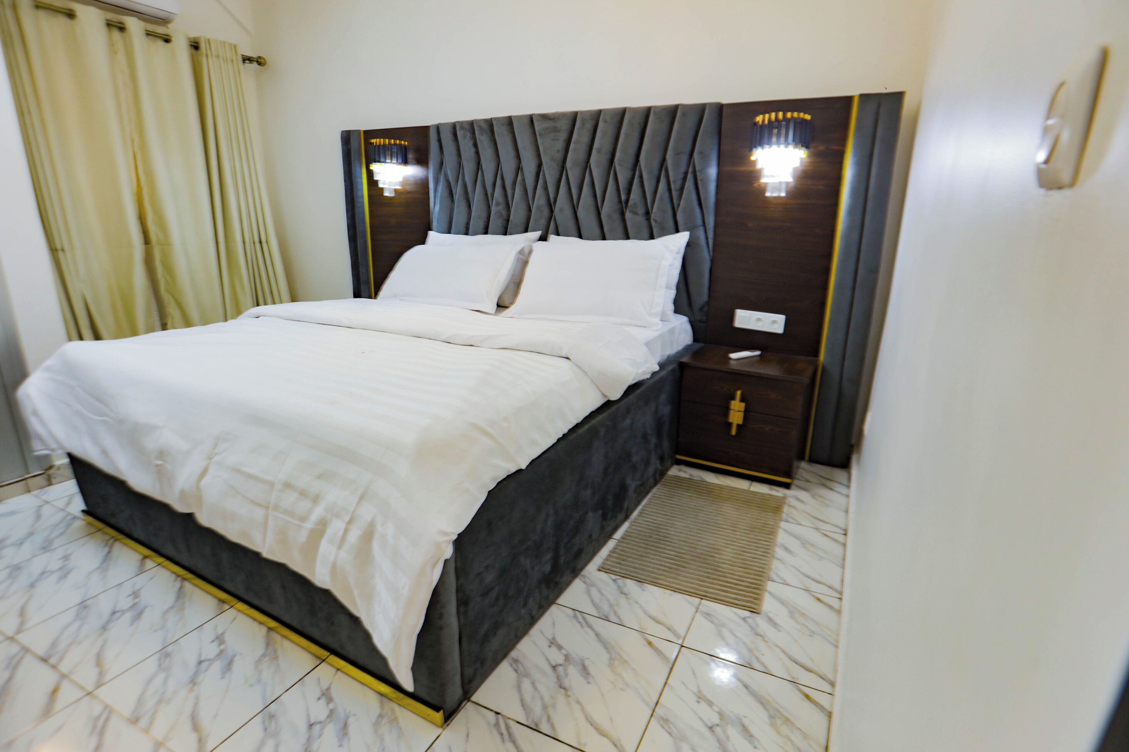 A neatly made bed with a dark headboard, a bedside table with a lamp, and a marble-tiled floor.