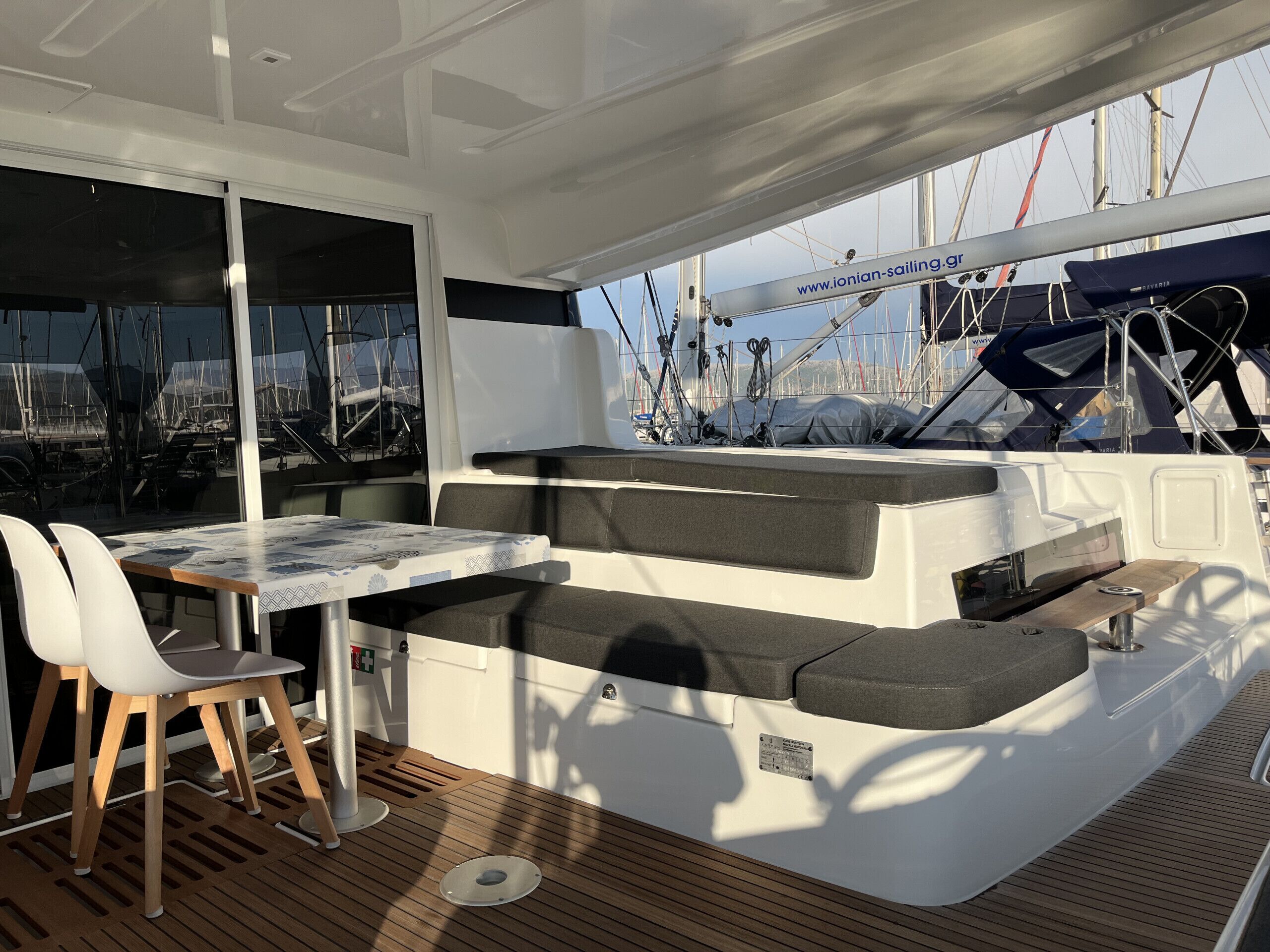 A sunlit deck with a table and chairs, a cushioned seating area, and a view of sailboats.