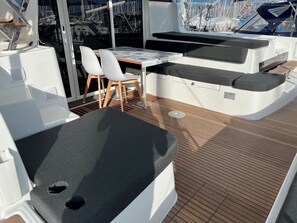 A sunlit deck with a table and chairs, a cushioned seating area, and a view of boats through a glass window. - Sun-Kissed Sailing Adventure in the Ionian Islands, Lefkada (Lefkada)
