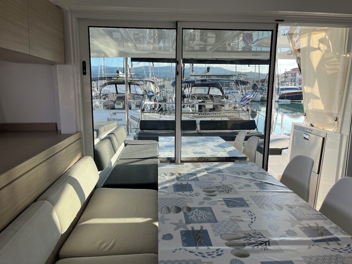 A boat interior with a dining area, cushioned seating, and a view of a marina with boats.