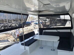 Interior de un barco con mesa, bancos, timón y vista al puerto deportivo.