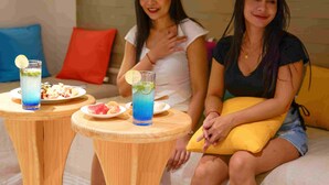 Twee vrouwen zitten aan een houten tafel met drankjes en eten, in een kamer met een bed en kussens.