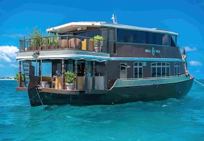 A large boat with a dining area, outdoor seating, and a name 'Atoll Villa' on the side.