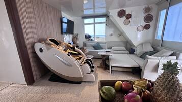 Una sala moderna con un sillón de masajes, un conjunto de sofás, una mesita y una cesta de frutas.