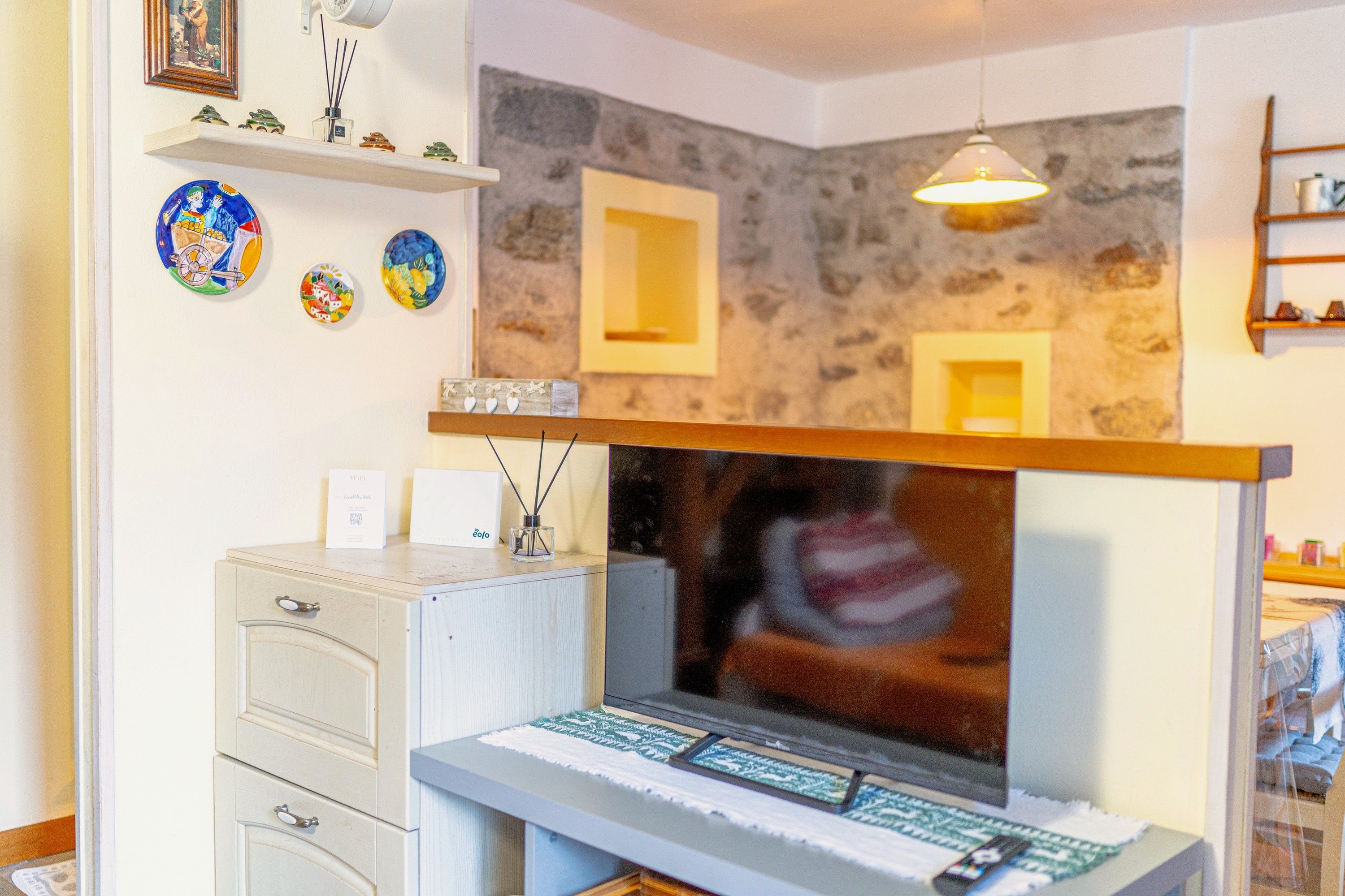 A room with a stone wall, a mounted television, a desk with a computer, and a shelf with decorative items.