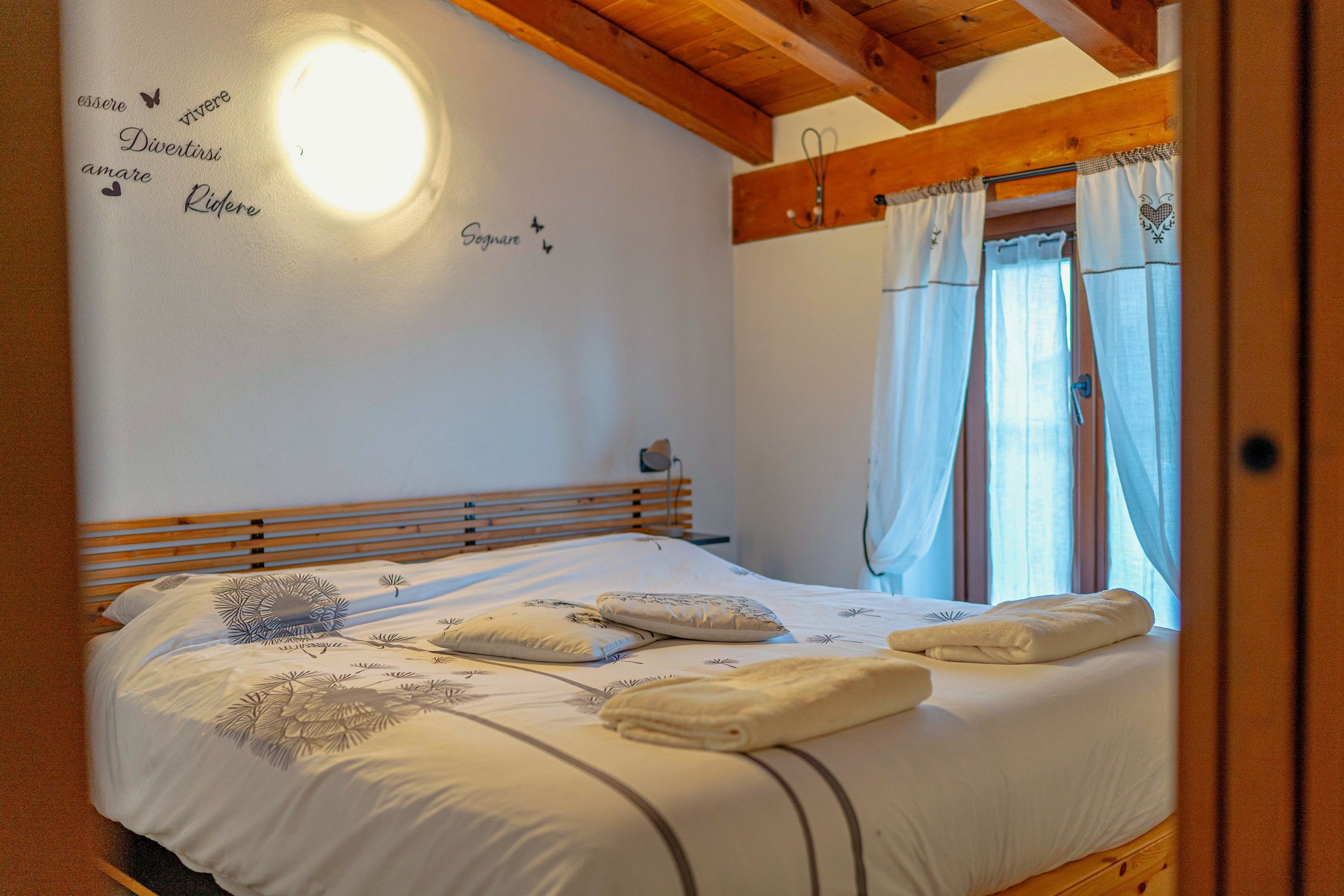Een slaapkamer met een houten hoofdeinde, een bed met witte beddengoed en decoratieve kussens, een wand met een lamp en een raam met gordijnen.