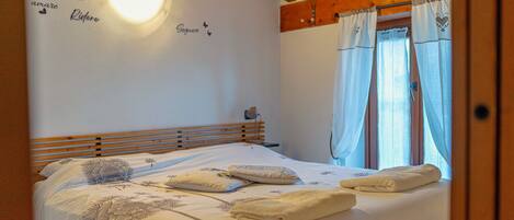 Un dormitorio con cabecera de madera, cama con ropa blanca y almohadas decorativas, una pared con una lámpara y una ventana con cortinas.