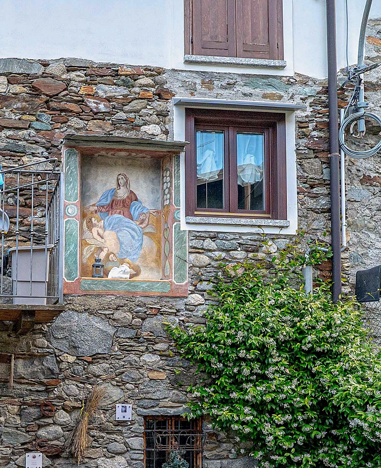Ein Steinhaus mit Wandgemälde, einem Fenster mit Rollläden und einem Metallgeländer.