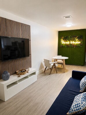 Ein modernes Wohnzimmer mit einem Flachbildfernseher, einer Essecke mit einem weißen Tisch und Stühlen sowie einer grünen Wand mit einer Pflanze.