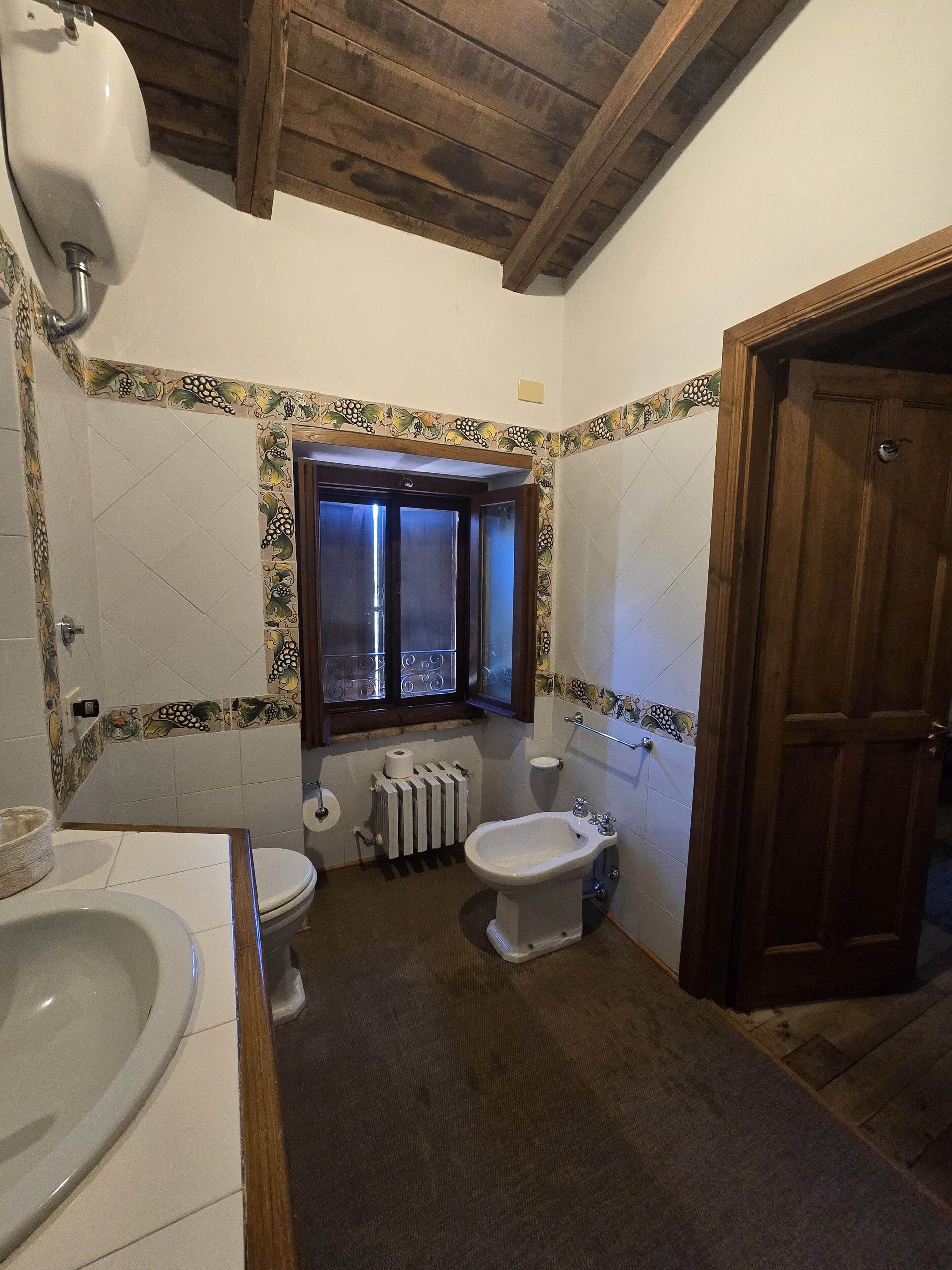 Ein Badezimmer mit Toilette, Bidet und einer Badewanne. Ein Fenster mit Vorhängen, ein Heizkörper und eine Holztür sind ebenfalls vorhanden.