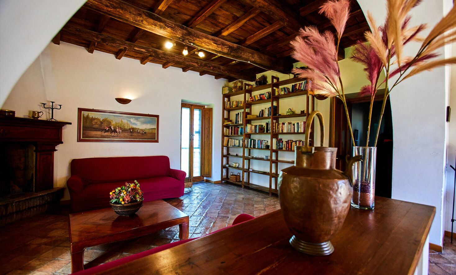 A cozy living room with a red sofa, a wooden coffee table, a bookshelf, and a large vase with pampas grass.