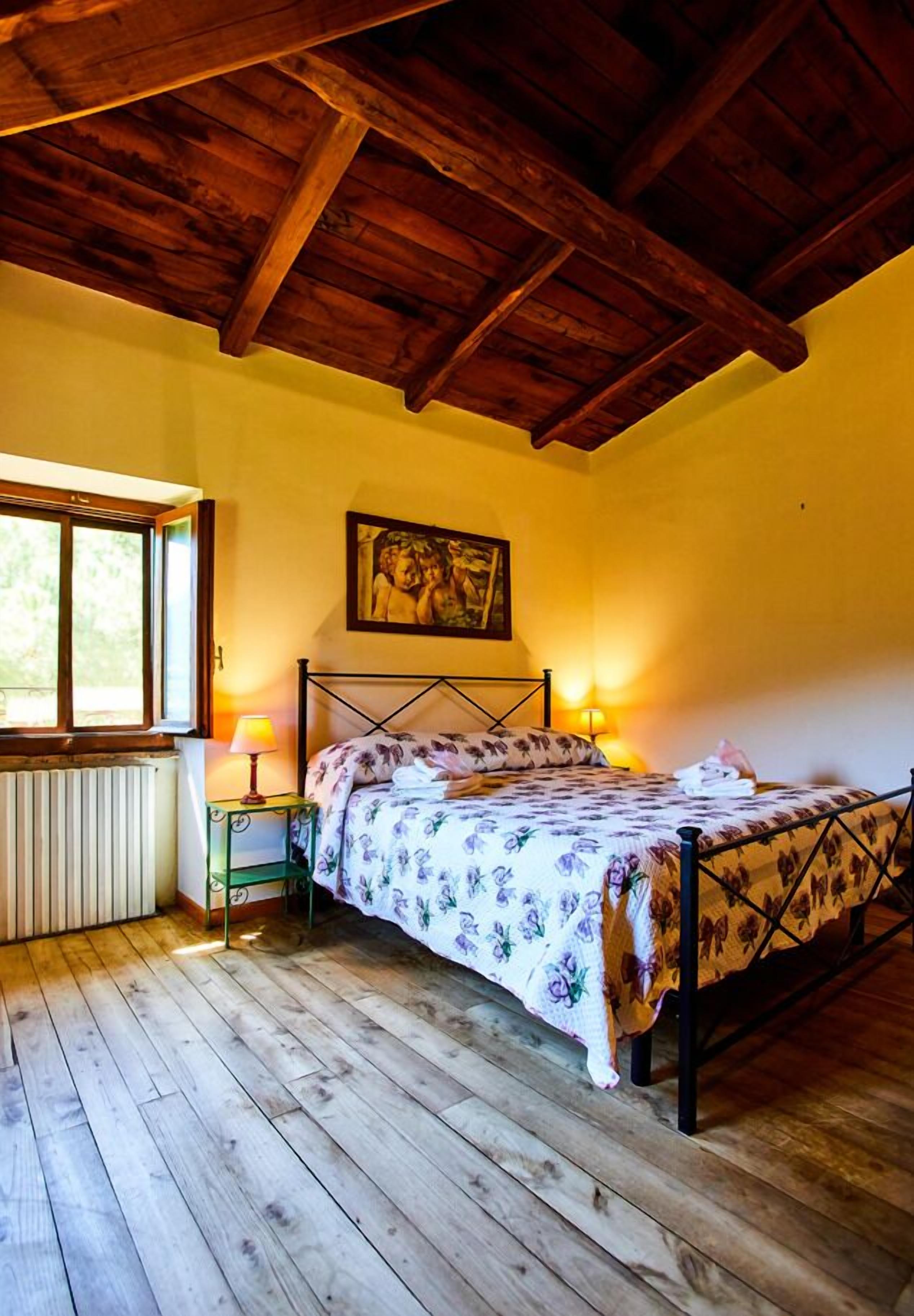 A bedroom with a wooden ceiling, a bed with a floral bedspread, a nightstand with a lamp, and a window providing natural light.