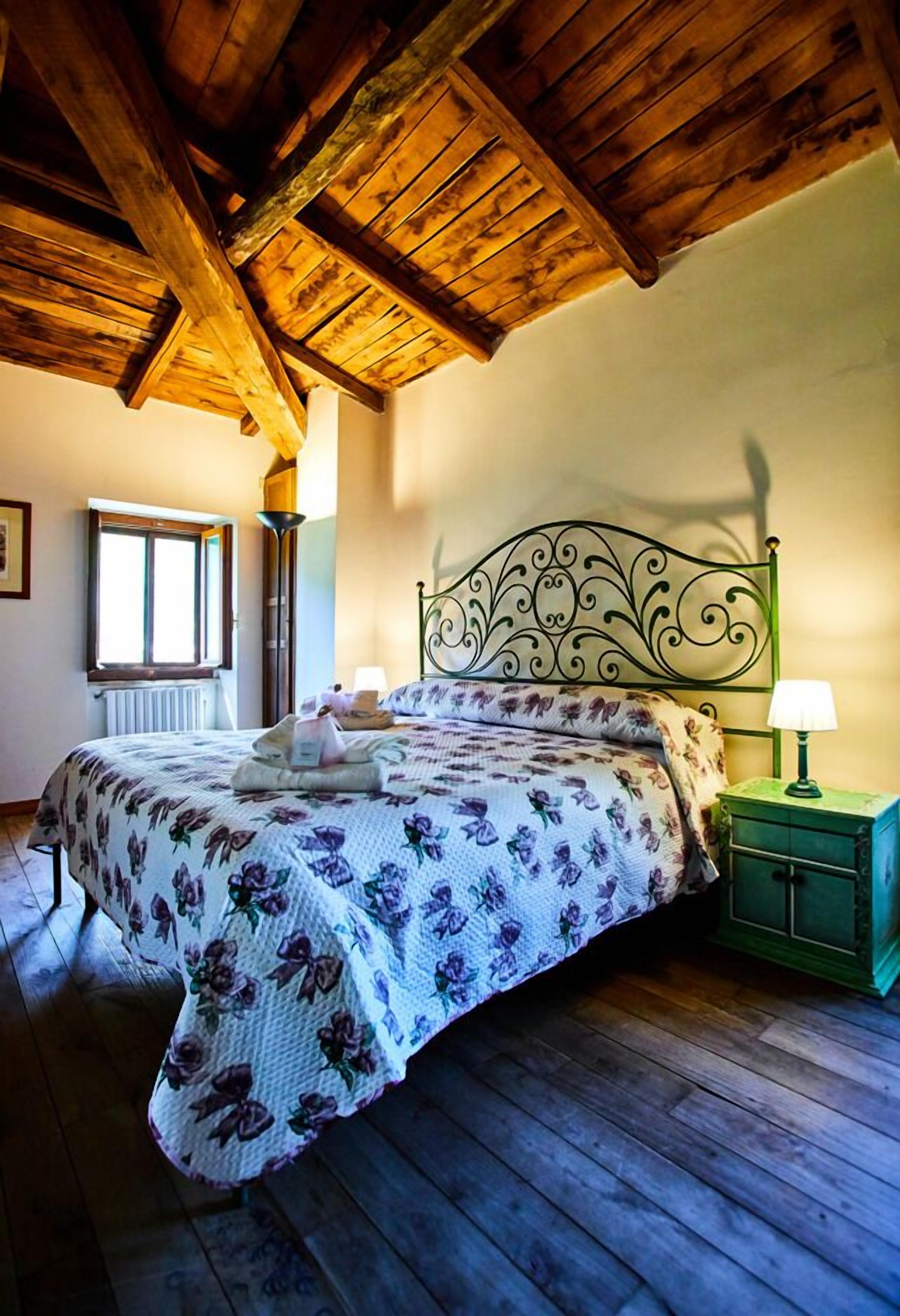 Une chambre avec un plafond en bois, un lit recouvert d’une couvre-lit à motifs floraux, une table de chevet avec une lampe et une fenêtre avec un radiateur.