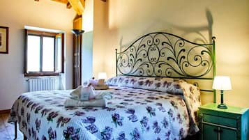 Une chambre avec un plafond en bois, un lit recouvert d’une couvre-lit à motifs floraux, une table de chevet avec une lampe et une fenêtre avec un radiateur.