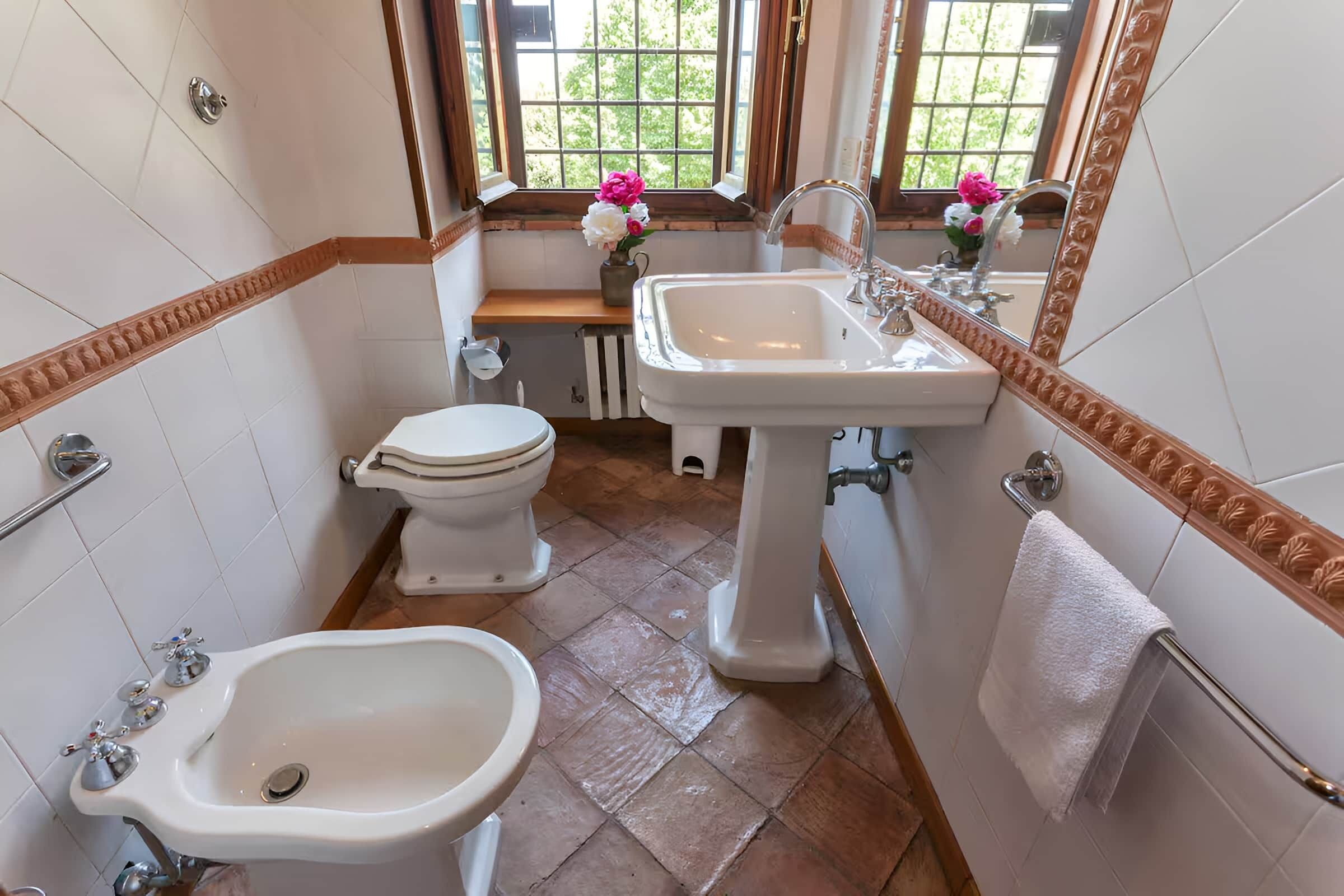 Ein Badezimmer mit Bidet, Toilette, Waschbecken und einem Fenster mit Blumen.