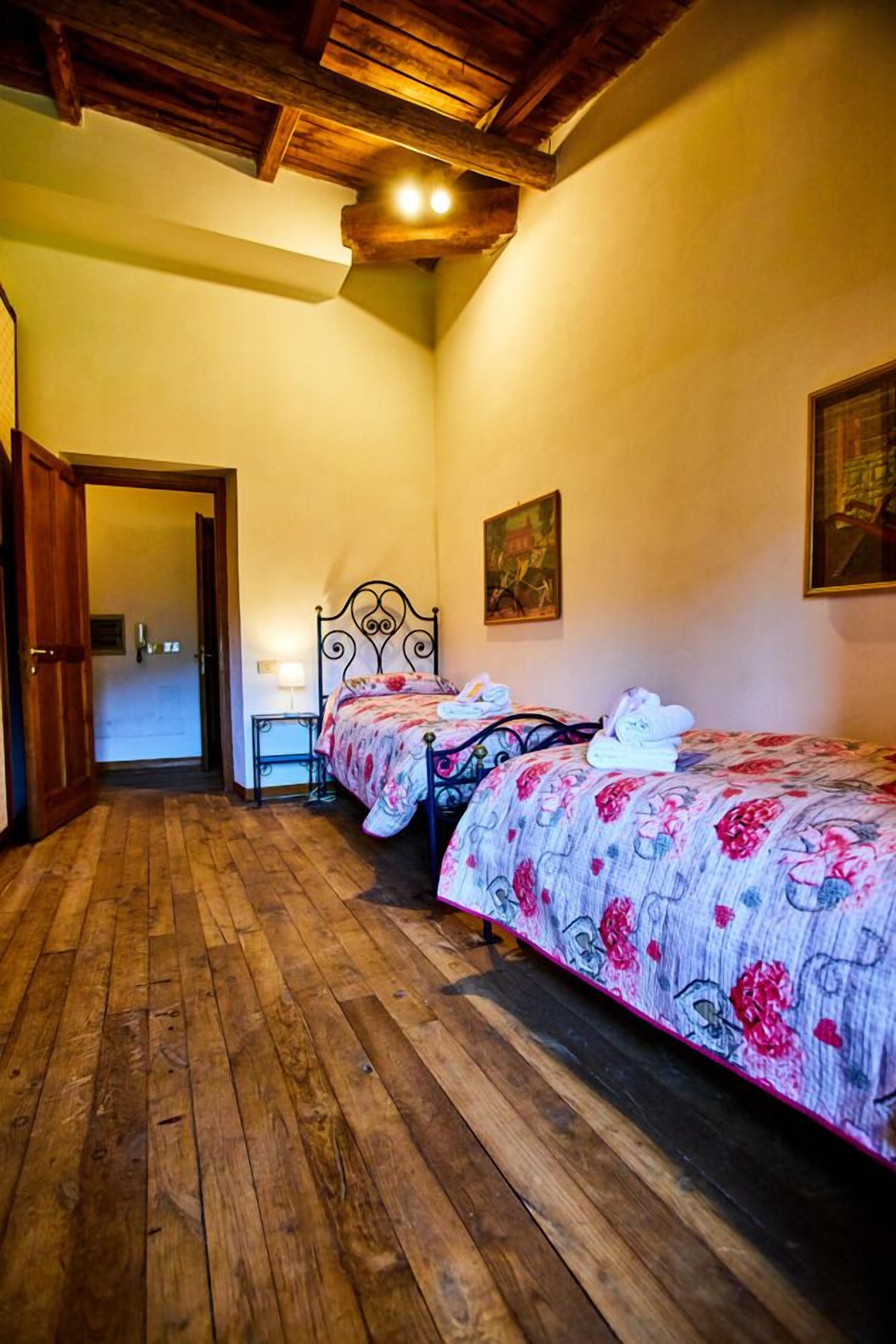 A bedroom with two beds, wooden flooring, a door, a nightstand, and a painting on the wall.