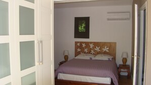 Quarto com cama, duas mesas de cabeceira, ar condicionado fixado na parede e janela com vista para o verde.