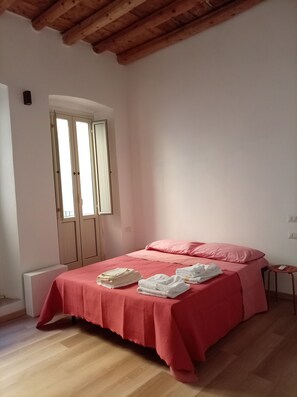 Ein Schlafzimmer mit Holzbalkendecke, einem Bett mit roter Bettdecke, einem rosafarbenen Kopfkissen und einem kleinen Hocker mit Fernbedienung.