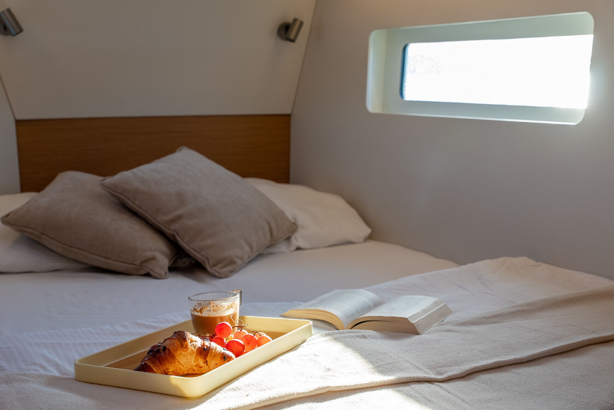 Een opgemaakt bed met kussens, een dienblad met gebakjes en een kopje koffie, en een open boek op een witte spreidek.