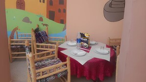 Un comedor con una mesa lista para dos personas, un mural en la pared y una silla de madera con un cojín colorido.