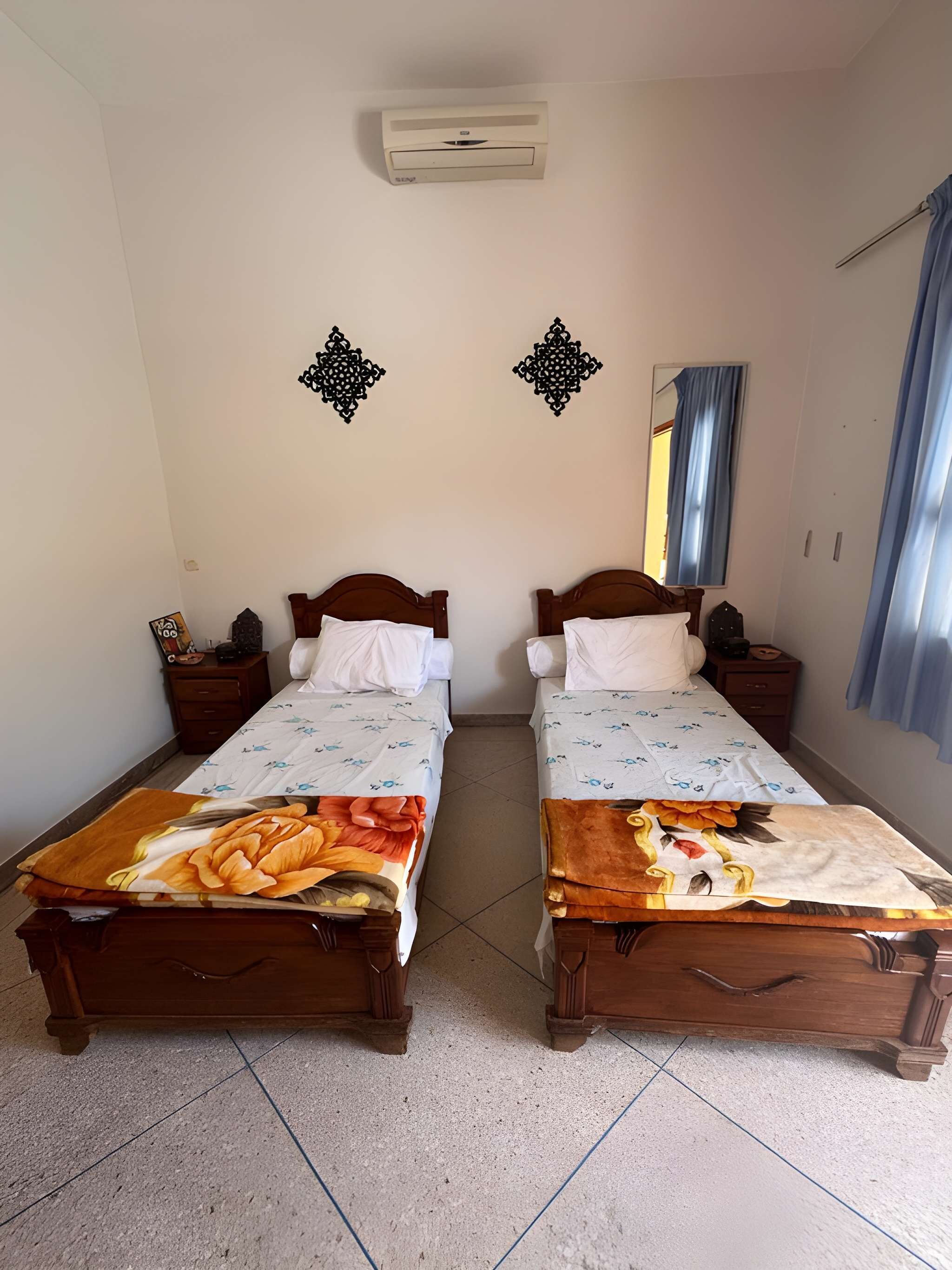 A room with two wooden beds, a wall-mounted air conditioner, decorative wall art, a mirror, and a window with curtains.