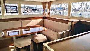 Schiffsinnere mit gepolsterten Sitzgelegenheiten, einem Holztisch und Fenstern mit Blick auf eine Marina.