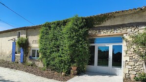 Un edificio con muros de piedra, un gran arbusto verde y una puerta con paneles de vidrio.