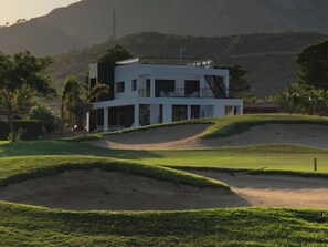 Ein modernes, zweistöckiges Gebäude mit flachem Dach und großen Fenstern, umgeben von einem Golfplatz mit Sandbunkern und üppiger Vegetation.