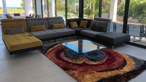 Ein modernes Wohnzimmer mit einem Ecksofa, einem Glastisch und großen Fenstern mit Blick ins Freie.