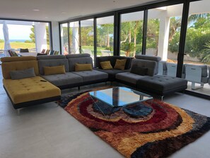 A modern living room with a sectional sofa, a glass coffee table, and large windows offering a view of the outdoors. - Golf course villa sleeping 8–10 with ocean views, 8 minutes from the beaches (Bonmont Terres Noves)