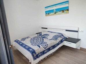 A bedroom with a bed, a nightstand, a painting of a beach scene, and a door. - Golf course villa sleeping 8–10 with ocean views, 8 minutes from the beaches (Bonmont Terres Noves)