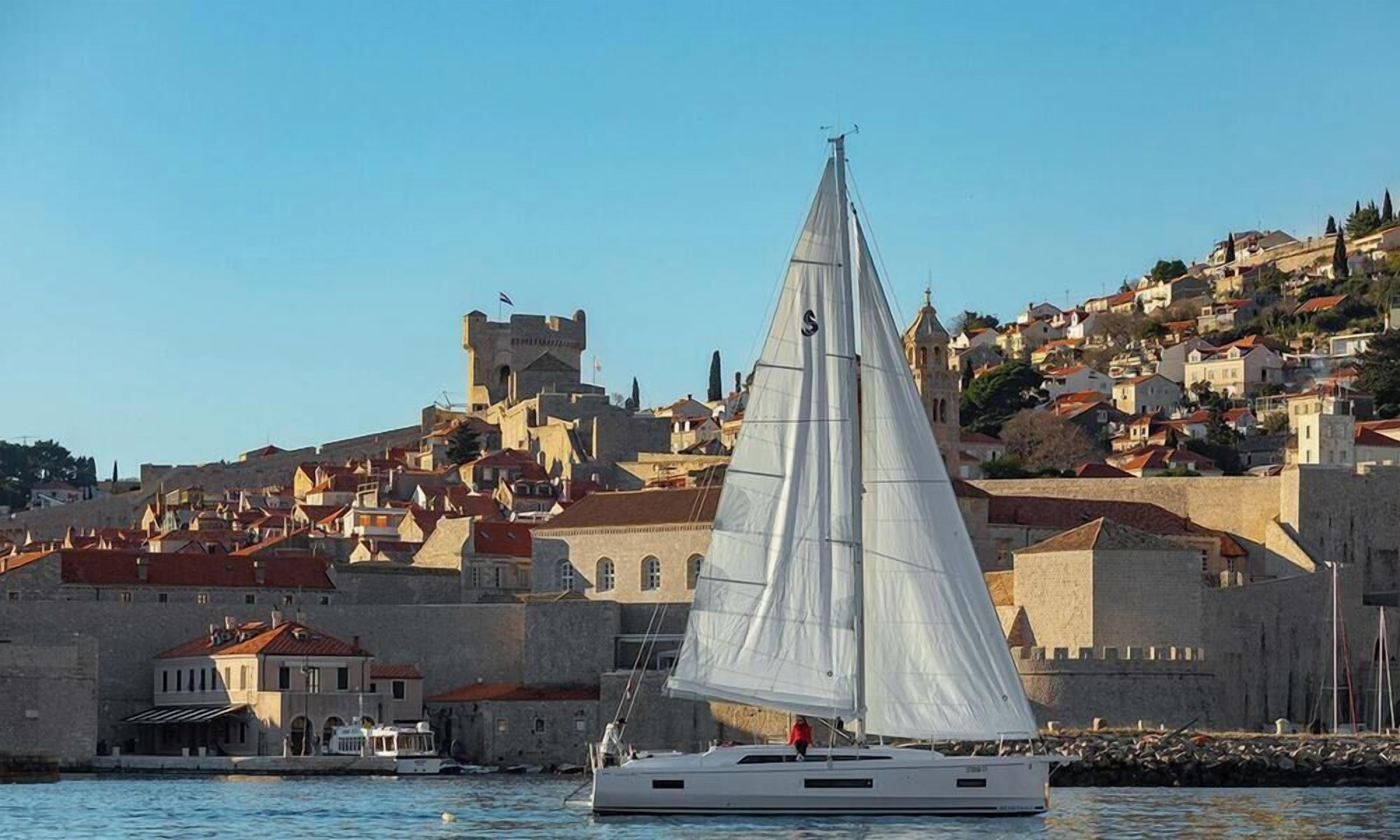 Un velero con vela blanca sobre aguas tranquilas, una pared de piedra histórica con tejados de tejas rojas y una colina con edificios al fondo.