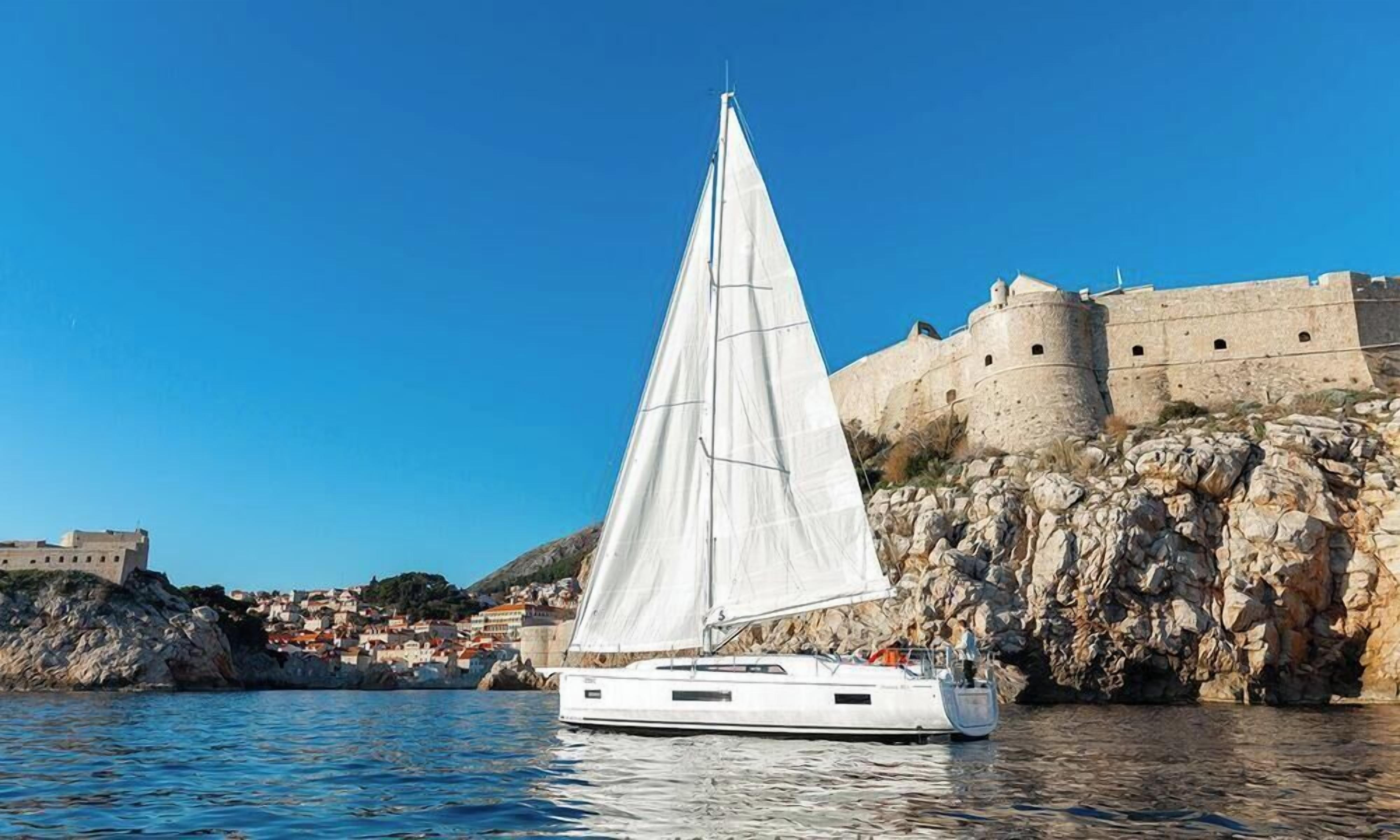 A sailboat with a white sail anchored near a rocky coastline with a historic fortification in the background.