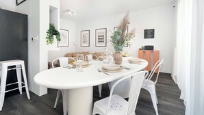 A modern dining area with a white table set for four, surrounded by white chairs. There are framed pictures on the wall, a potted plant, and a vase with dried plants. - Unique 2-Bedroom Apartment Balcony Parking (Poznań)
