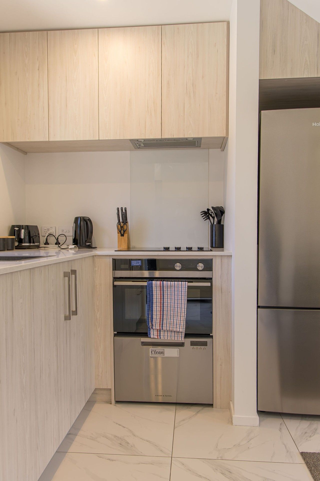 A modern kitchen with light wood cabinetry, stainless steel appliances including a refrigerator, oven, and coffee maker, and a tiled backsplash.