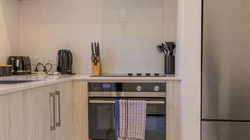 A modern kitchen with light wood cabinetry, stainless steel appliances including a refrigerator, oven, and coffee maker, and a tiled backsplash.