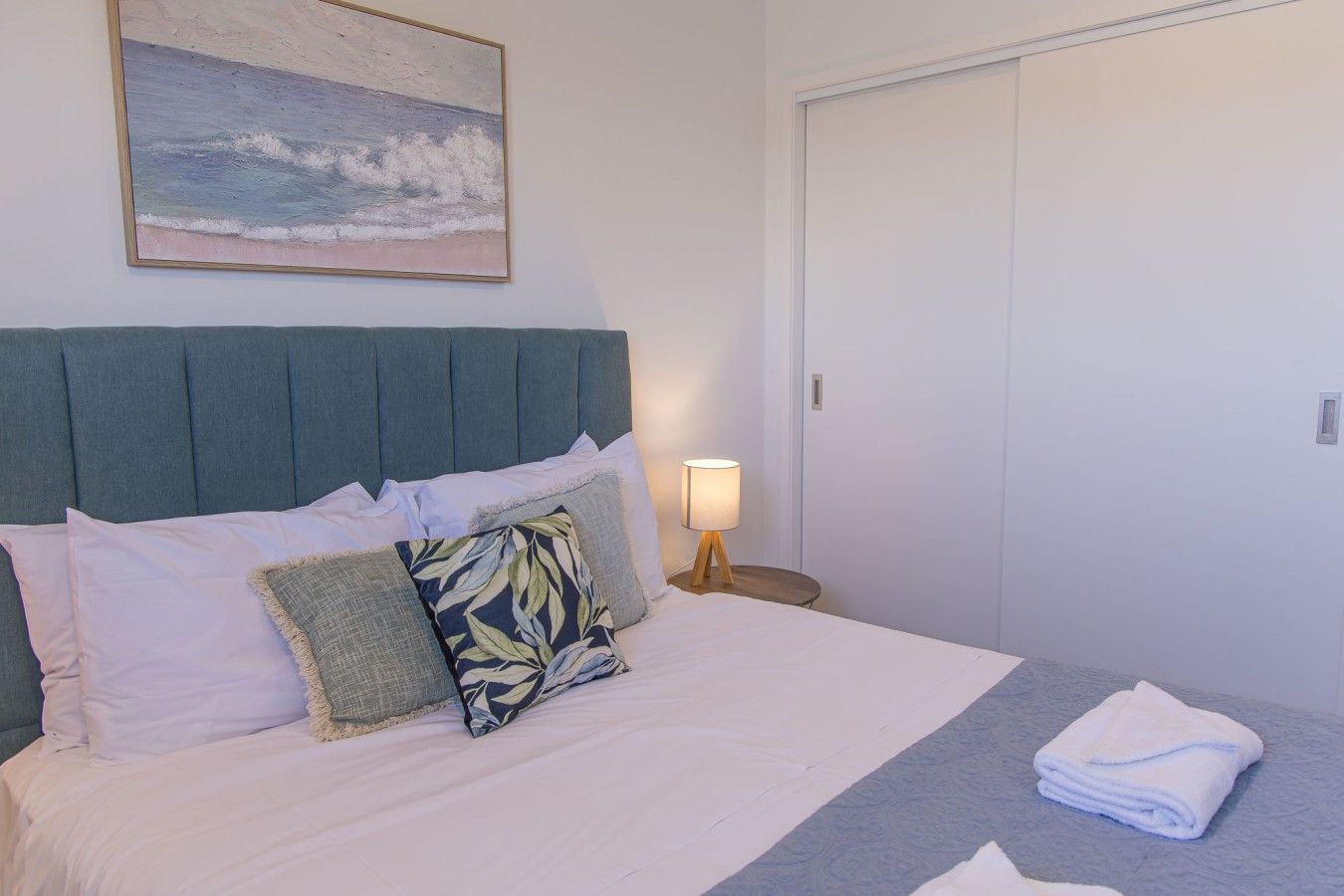 A neatly made bed with pillows and a folded towel, a bedside lamp, and a painting of a beach scene on the wall.