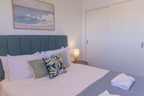 A neatly made bed with pillows and a folded towel, a bedside lamp, and a painting of a beach scene on the wall.