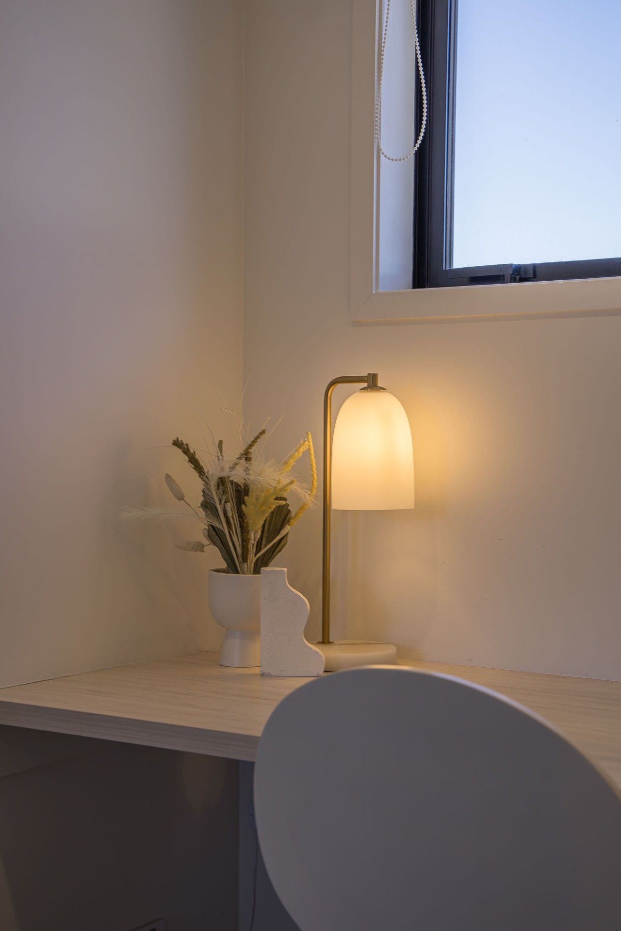 Ein schlichter Schreibtisch mit weißem Stuhl, einer Vase mit Trockenpflanzen und einer Wandlampe.