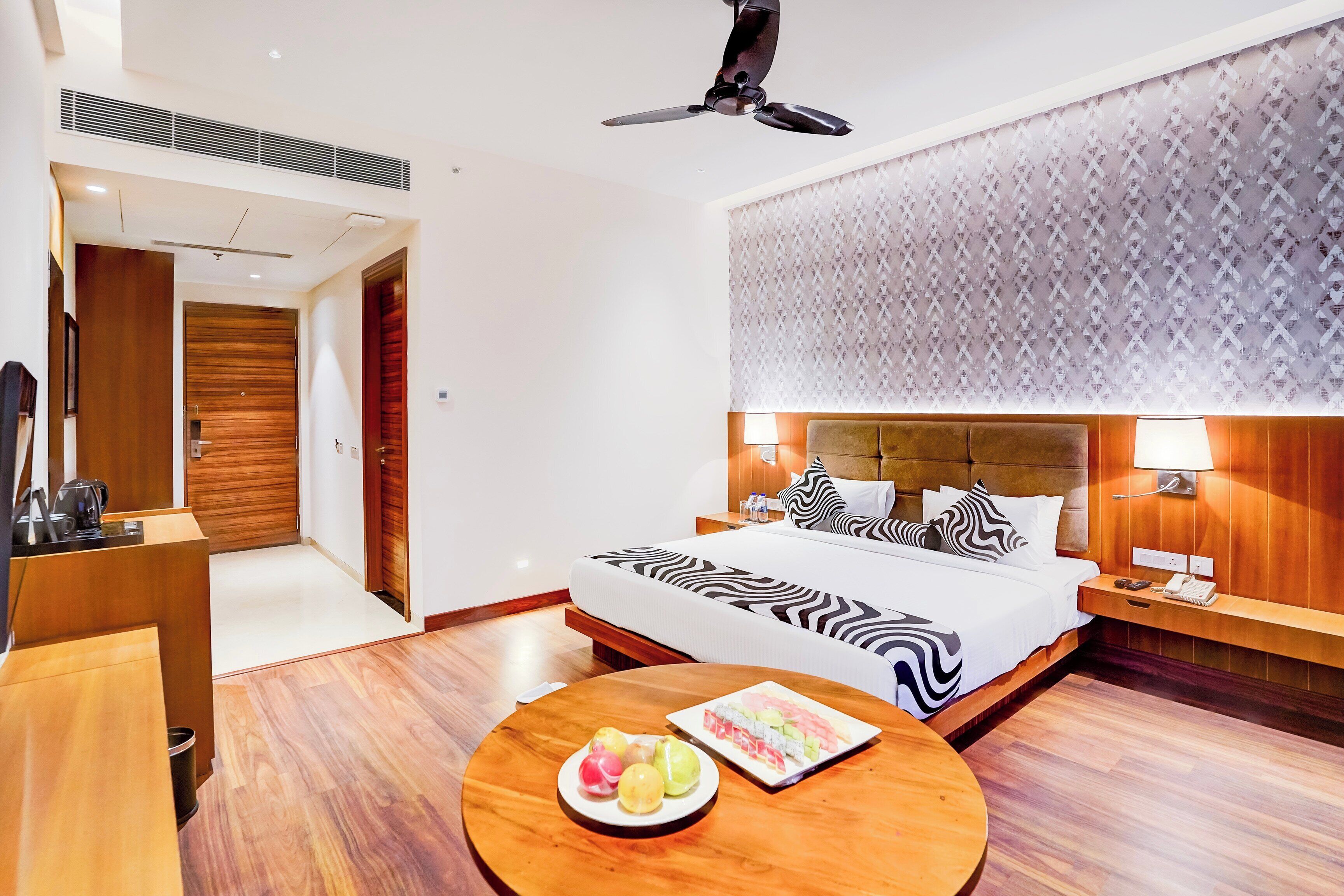 Une chambre d’hôtel avec un lit, des tables de chevet, une table ronde en bois sur laquelle sont disposés des fruits, un ventilateur de plafond et un mur décoré d’un motif.