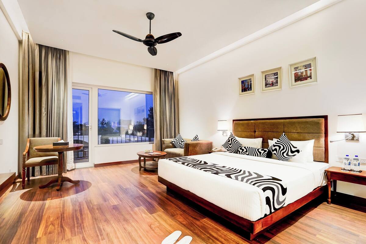 Una habitación de hotel con una cama grande, ventilador de techo, vista por la ventana y un área de descanso con mesa y sillas.