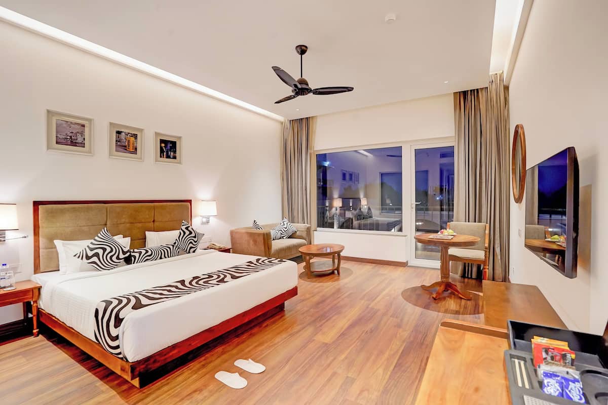 Habitación de hotel con una cama grande, ventilador de techo, televisor, una mesita y una ventana con cortinas.