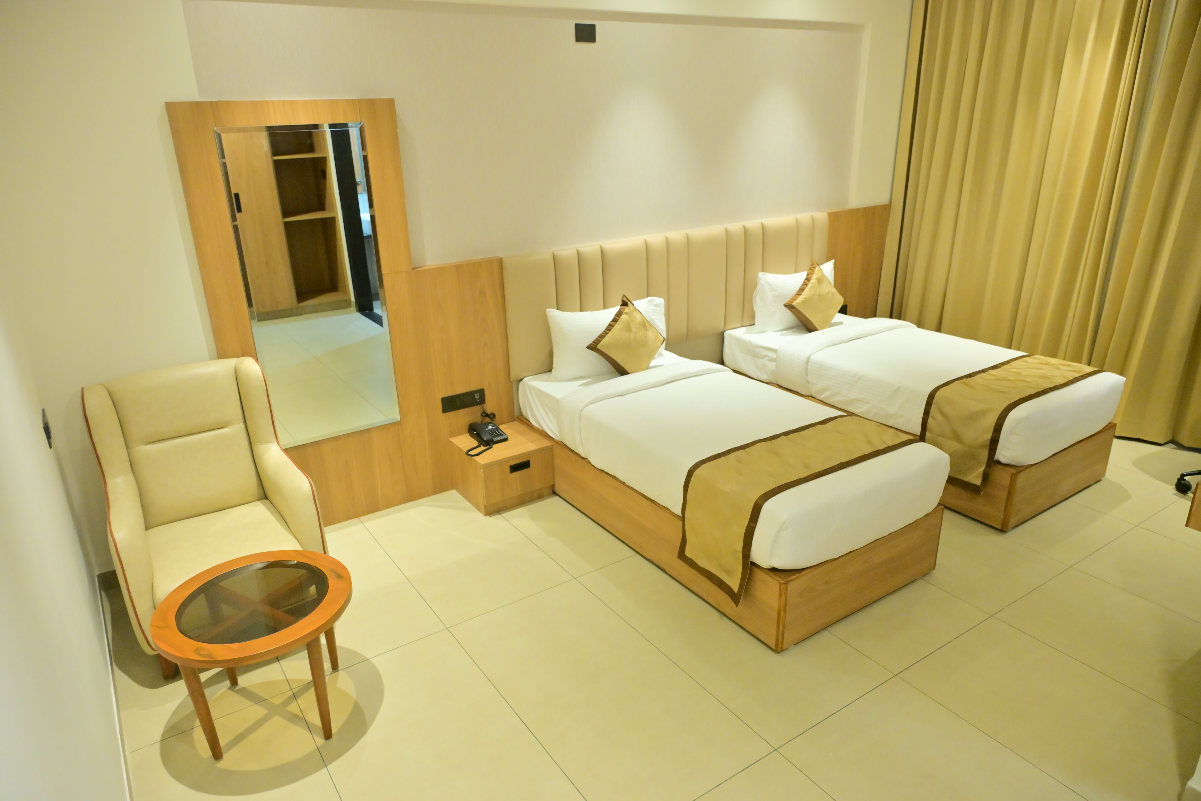 A hotel room with two beds, a chair, a small table, a bedside table with a phone, and a mirror.